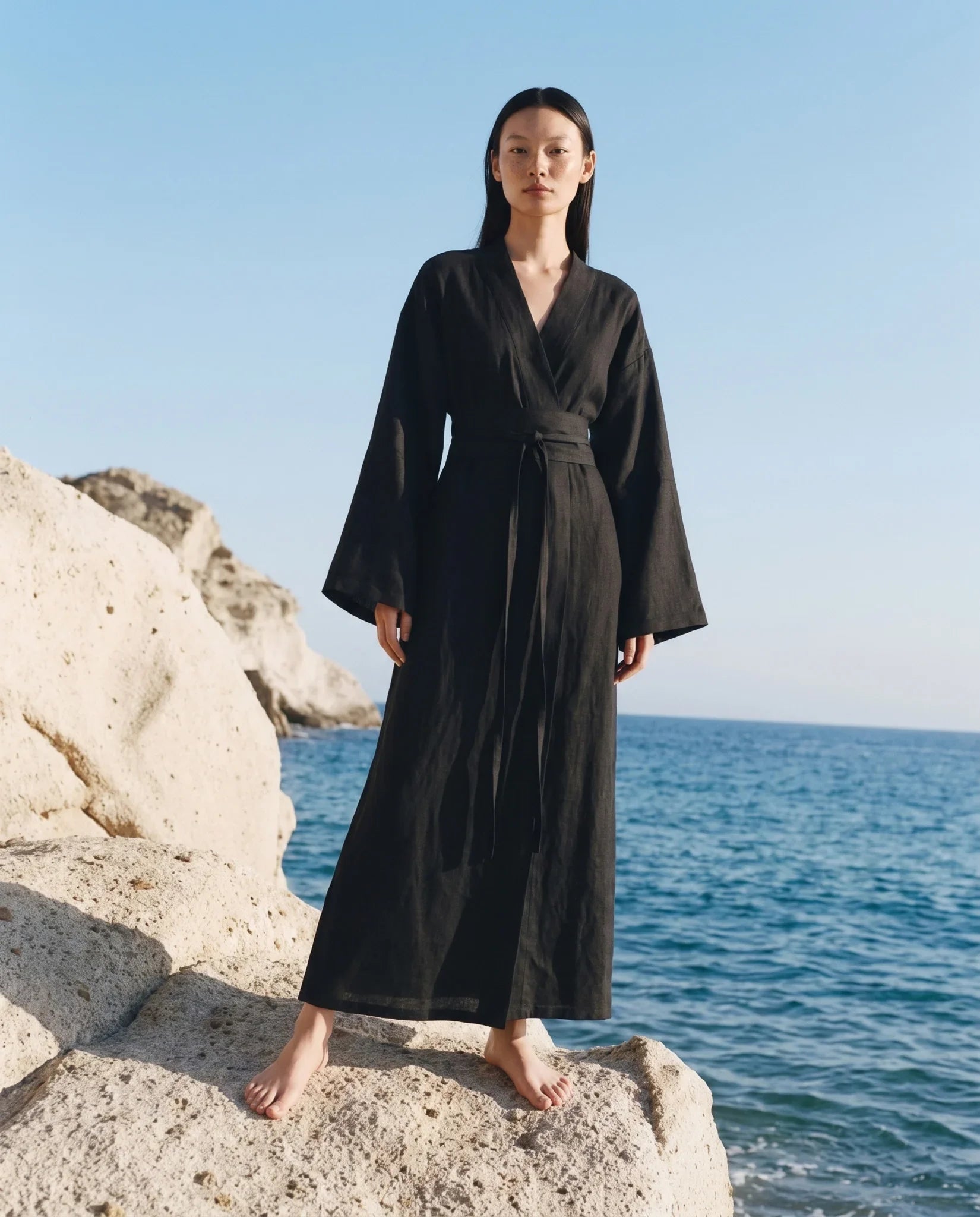 A woman stands barefoot on a rocky shoreline wearing a long black wrap-style maxi dress with wide sleeves and a self-tie belt, with the blue sea and sky behind her.